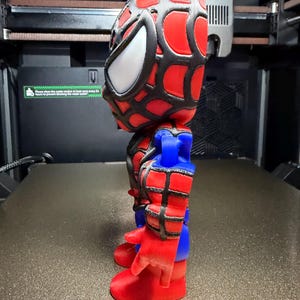 May include: A 3D-printed Spider-Man figurine. The figure has a red and black webbed mask with white eye lenses, red and blue suit, and red gloves and boots. The figurine is standing on a textured surface.
