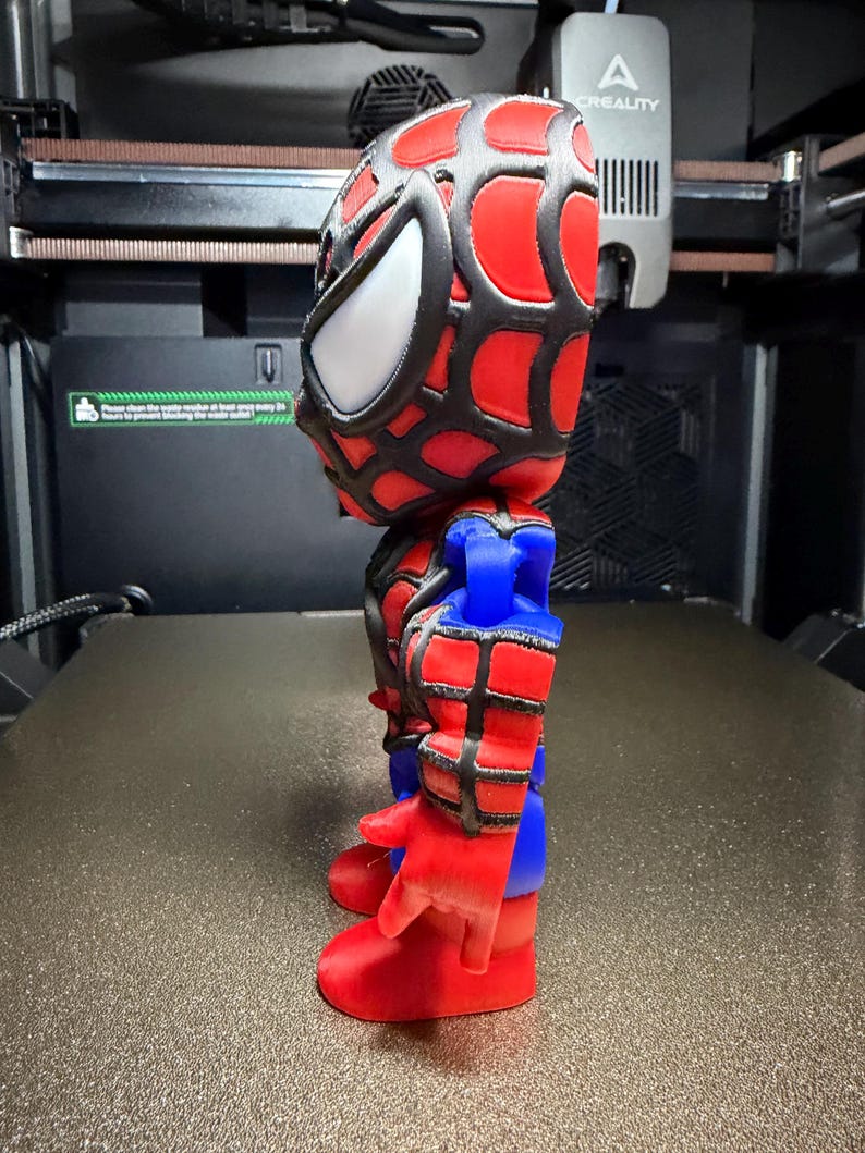 May include: A 3D-printed Spider-Man figurine. The figure has a red and black webbed mask with white eye lenses, red and blue suit, and red gloves and boots. The figurine is standing on a textured surface.