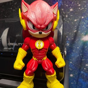 May include: A red and yellow action figure, a fusion of The Flash and Sonic the Hedgehog. The figure has a red head with yellow ears, a yellow lightning bolt emblem, and yellow gloves and boots. The figure is standing upright.