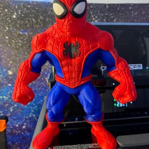 K&ouml;nnte beinhalten: Eine rot, blau und schwarz gehaltene Spider-Man-Actionfigur. Die Figur hat ein rotes und schwarzes Spinnenemblem auf der Brust. Die Figur steht mit ausgestreckten Armen da. Sie hat wei&szlig;e Augen und eine rote Maske.