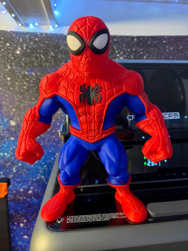 K&ouml;nnte beinhalten: Eine rot, blau und schwarz gehaltene Spider-Man-Actionfigur. Die Figur hat ein rotes und schwarzes Spinnenemblem auf der Brust. Die Figur steht mit ausgestreckten Armen da. Sie hat wei&szlig;e Augen und eine rote Maske.