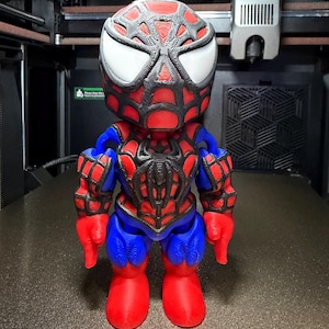 May include: A 3D-printed Spider-Man figurine. The figure is predominantly red and blue, with black detailing. The head has large white eyes and a webbed pattern. The figurine is standing on a textured surface.