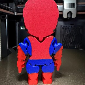 May include: A 3D-printed figurine, seen from behind, with a red head and torso, blue arms and legs, and red hands and feet. The figure is on a textured surface, with a 3D printer in the background.