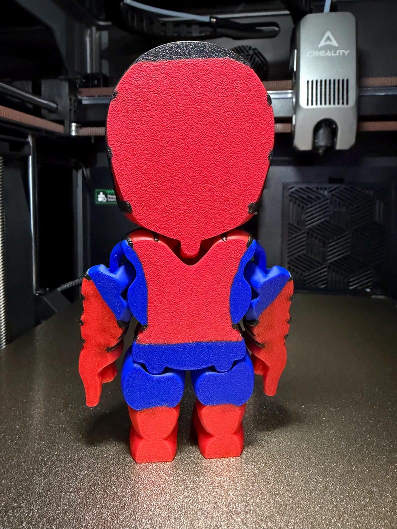 May include: A 3D-printed figurine, seen from behind, with a red head and torso, blue arms and legs, and red hands and feet. The figure is on a textured surface, with a 3D printer in the background.