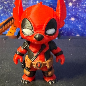 Puede incluir: Figura impresa en 3D de Stitch con un disfraz de Deadpool. La figura es principalmente roja con detalles en negro, incluyendo los ojos y la máscara. Cuenta con un cinturón de herramientas y un diseño detallado.