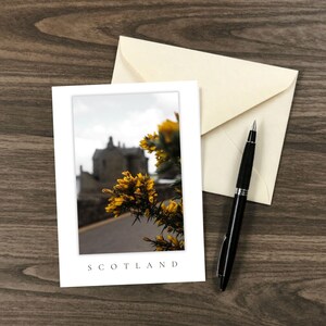 Könnte beinhalten: Eine weiße Grußkarte mit einem Foto von gelben Ginsterblüten im Vordergrund, einem unscharfen Steingebäude im Hintergrund und dem Wort SCOTLAND am unteren Rand, dazu ein cremefarbener Umschlag und ein schwarzer Kugelschreiber.