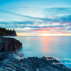 May include: Scenic landscape photograph of a coastal scene at dusk. The image features a lighthouse on a cliff, with a calm sea and a colorful sky. The foreground shows dark, rocky formations.