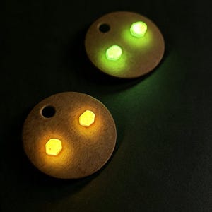 May include: Two circular, flat, tan-colored discs, each featuring a single circular hole near the top edge and two small, glowing hexagonal inserts. One disc displays bright green glowing hexagons, while the other disc displays warm yellow glowing hexagons.