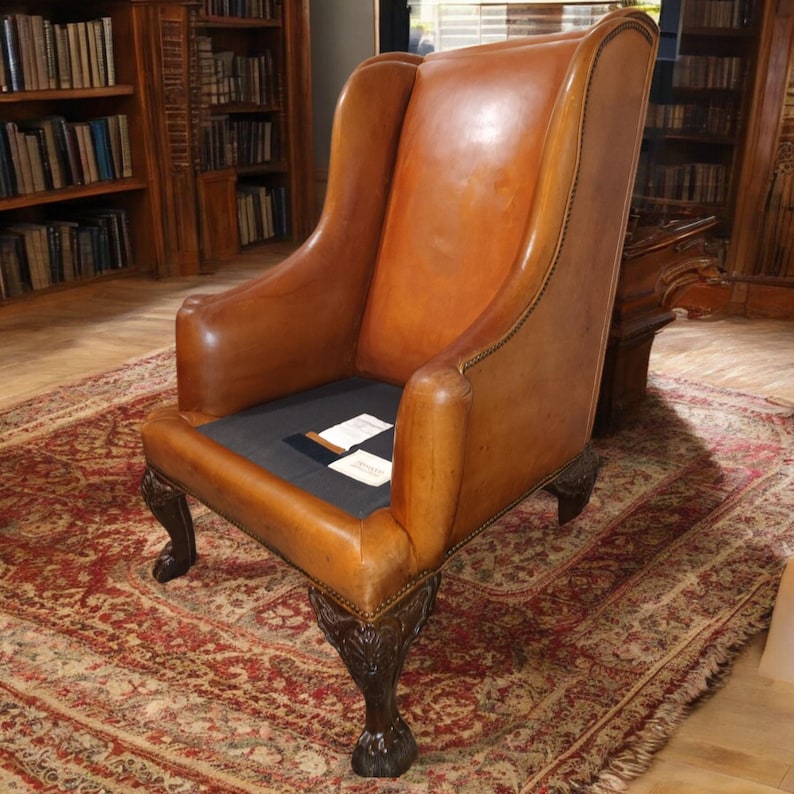 Op de afbeelding: Een vintage leren fauteuil in een warme bruine tint. De stoel heeft een wingback-ontwerp, gerolde armen en sierlijke, donkere houten poten. De zitting is van donkere stof. De stoel staat op een patroonkleed.