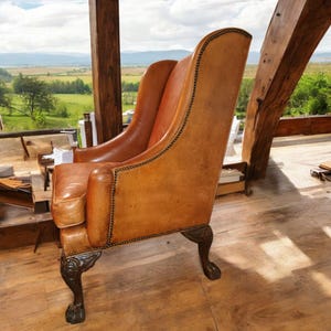 Op de afbeelding: Een bruin lederen fauteuil met een hoge rug en sierlijke donkere houten poten. De stoel heeft een geplooide rand en staat bij een raam met uitzicht op een groen landschap. Het ontwerp suggereert een klassieke, vintage stijl.