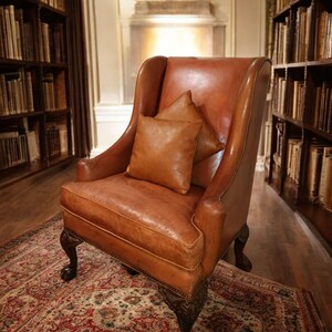 Op de afbeelding: Een klassieke bruin lederen fauteuil met een hoge rugleuning en twee bijpassende kussens. De stoel staat in een bibliotheek met boekenkasten vol boeken. De stoel staat op een patroonkleed, wat een warme sfeer cre&euml;ert.