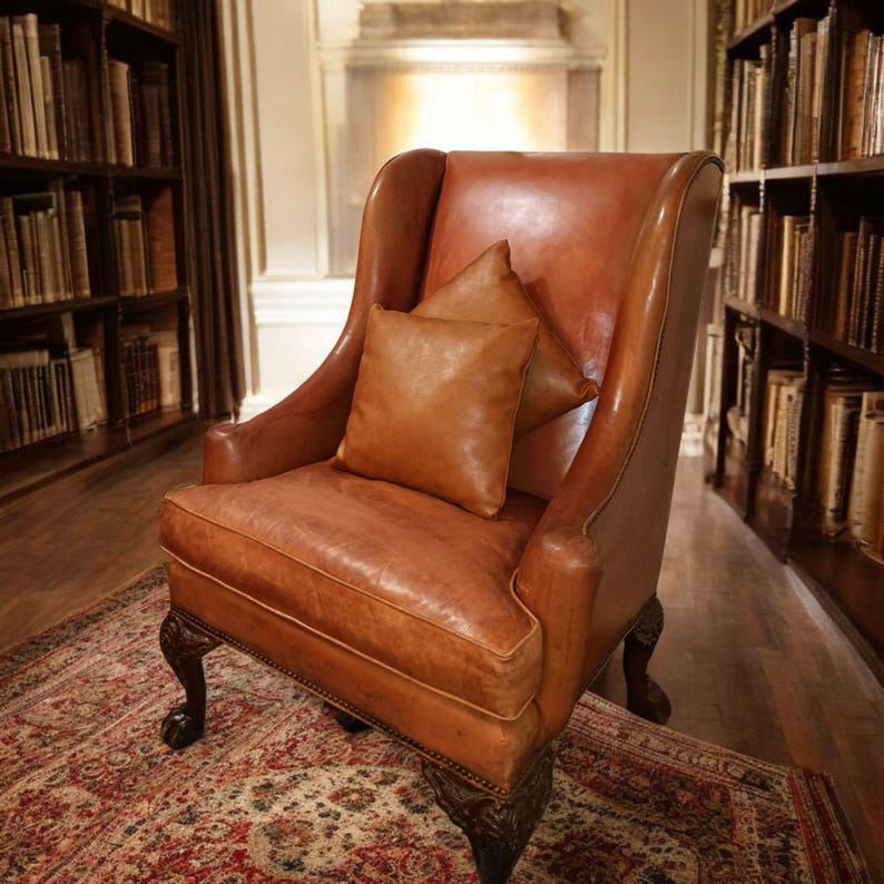 Op de afbeelding: Een klassieke bruin lederen fauteuil met een hoge rugleuning en twee bijpassende kussens. De stoel staat in een bibliotheek met boekenkasten vol boeken. De stoel staat op een patroonkleed, wat een warme sfeer cre&euml;ert.