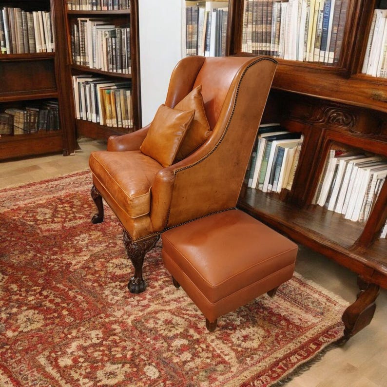Op de afbeelding: Een bruine leren fauteuil met bijpassende voetenbank en twee kussens. De stoel heeft een hoge rugleuning en sierlijke houten poten. De set staat op een rood tapijt met patronen, met boekenkasten op de achtergrond.