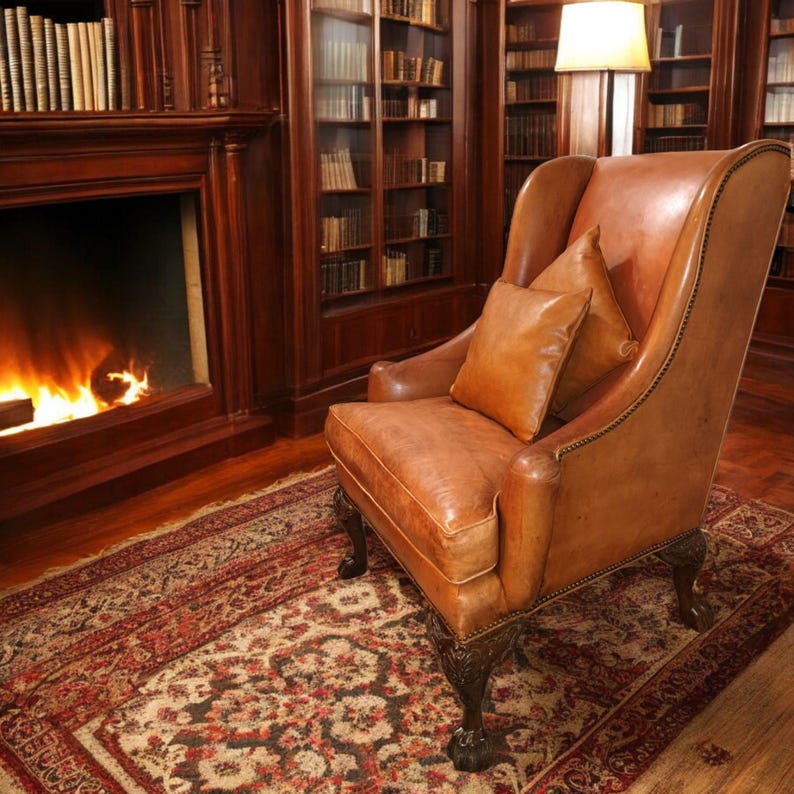Op de afbeelding: Een bruine leren fauteuil met kussens in een bibliotheek. De stoel staat op een patroonkleed, met een open haard en boekenkasten op de achtergrond. De kamer heeft een warme, uitnodigende sfeer.