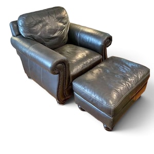 May include: A dark charcoal gray leather armchair with a matching rectangular ottoman. The chair features rolled arms, a thick cushioned backrest, and a seat cushion, all accented with decorative metal nailhead trim along the front edges.