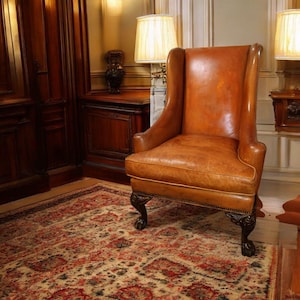 Op de afbeelding: Een vintage leren fauteuil met hoge rug in een kamer met een patroonkleed. De stoel heeft een warme bruine kleur met een verweerde uitstraling. De poten zijn donker en sierlijk. De kamer heeft lambrisering en lampen.