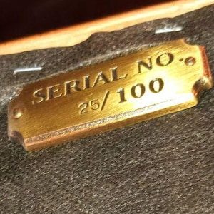 Op de afbeelding: Een rechthoekige messing plaat met de tekst "SERIAL NO." en "25/100" gegraveerd. De plaat heeft afgeronde hoeken en is geplaatst tegen een donkere, getextureerde achtergrond. De plaat heeft een warme goudkleur.