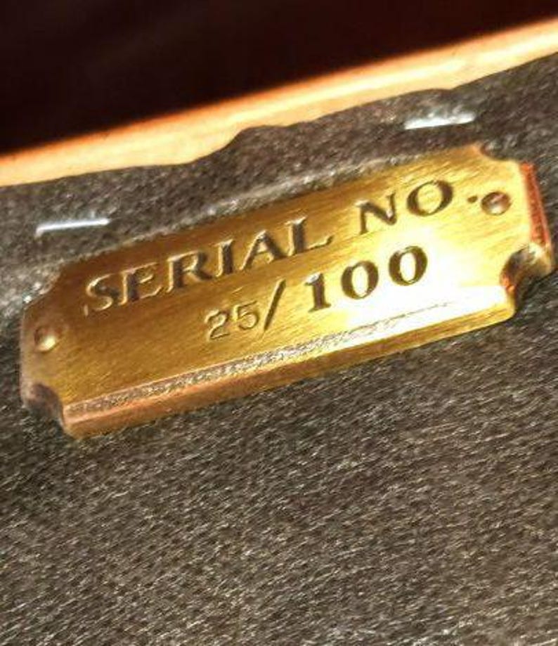 Op de afbeelding: Een rechthoekige messing plaat met de tekst "SERIAL NO." en "25/100" gegraveerd. De plaat heeft afgeronde hoeken en is geplaatst tegen een donkere, getextureerde achtergrond. De plaat heeft een warme goudkleur.