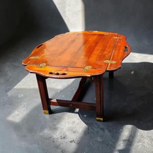 May include: A rectangular wooden tray table with a warm brown finish, featuring brass hinges and corner accents. The tabletop has integrated cutout handles on the sides and is supported by four straight legs with brass feet at the base.