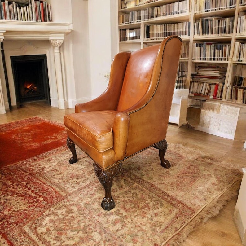 Op de afbeelding: Een vintage leren fauteuil in een warme bruine tint, met een hoge rugleuning en armleuningen. De stoel staat op een patroonkleed in een kamer met een open haard en boekenkasten, wat een klassieke en gezellige sfeer cre&euml;ert.