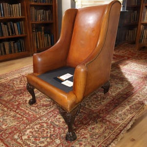 Op de afbeelding: Een vintage fauteuil met hoge rugleuning en bruin leer. De stoel heeft een wingback-ontwerp, gebogen armleuningen en donkere, sierlijke klauwpoten. Het zitkussen is donker. De stoel staat op een patroonkleed.