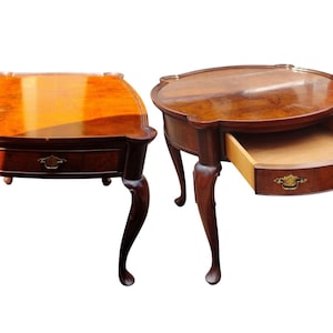 May include: Two brown wooden side tables with polished surfaces and curved legs. Each table features a single drawer with a decorative brass handle. The table on the left has a square top, while the table on the right has a rounded top.