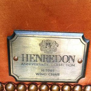 Op de afbeelding: Close-up van een label van een Henredon Anniversary Collection wing chair. Het messing label toont het Henredon-logo, tekst en het modelnummer H.1945. Het label is bevestigd aan een bruine leren stoel met decoratieve messing studs.