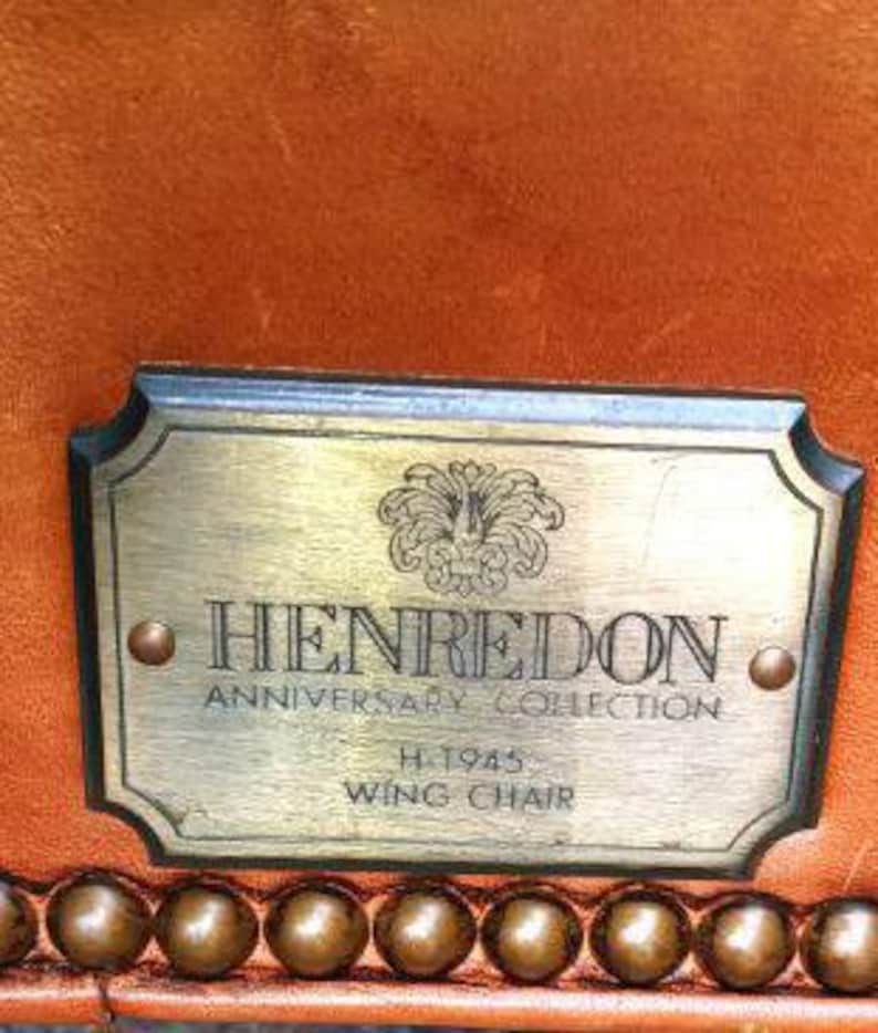 Op de afbeelding: Close-up van een label van een Henredon Anniversary Collection wing chair. Het messing label toont het Henredon-logo, tekst en het modelnummer H.1945. Het label is bevestigd aan een bruine leren stoel met decoratieve messing studs.