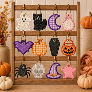 May include: A wooden display rack holding twelve embroidered keychains featuring Halloween motifs including a ghost, black cat, crescent moon, potion bottle, bat, pumpkin, coffin, jack-o'-lantern, spider, skull, witch hat, and a star.