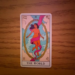 Könnte beinhalten: Eine Tarotkarte mit einer nackten Figur in einem Lorbeerkranz, mit einem rosa Band und zwei Stäben. Die Karte trägt unten die Aufschrift "DIE WELT", mit astrologischen Symbolen und Wolken im Hintergrund.