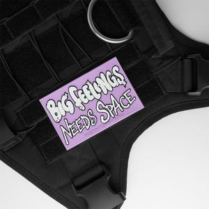 Op de afbeelding: Zwart hondentuig met een paarse en witte patch. De patch zegt "Big Feelings Needs Space". Het tuig heeft meerdere banden, een metalen ring en een plastic gesp. De tekst "mightyiga.com.au" staat onderaan de patch.