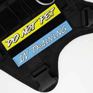 Peut inclure: Harnais noir pour chien avec deux écussons rectangulaires. Un écusson est jaune avec le texte "DO NOT PET" en blanc. L'autre écusson est bleu clair avec le texte "IN TRAINING" en blanc. Le harnais a une boucle noire et un anneau métallique.