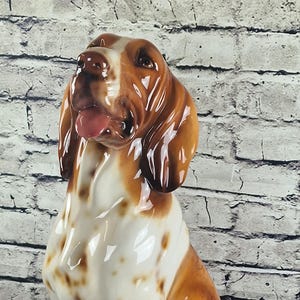 Pode incluir: Estatueta de cão em cerâmica, em posição sentada, com acabamento brilhante. O cão tem pelo manchado castanho e branco, com cabeça e orelhas castanhas. Sua língua é visível. Fundo: parede de tijolo e chão de madeira.