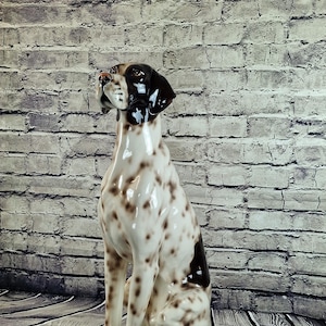 Puede incluir: Figura de perro de cerámica en posición sentada, con acabado brillante. El perro tiene una base blanca con manchas marrones y cabeza y orejas de color marrón oscuro. La figura está colocada contra una pared de ladrillos grises y blancos y un suelo de madera.