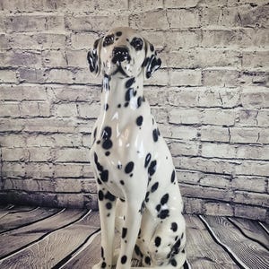 May include: A ceramic Dalmatian dog statue, featuring a glossy white finish with black spots. The dog is in a seated position, with its ears and nose detailed in black. The statue is a decorative item.