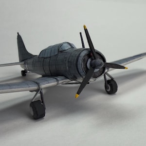 Puede incluir: Maqueta detallada de avión en tonos grises y negros, con una hélice con aspas con punta amarilla. El modelo tiene cabina y alas, y está sobre un fondo blanco. La envergadura del avión es de aproximadamente 25 cm.