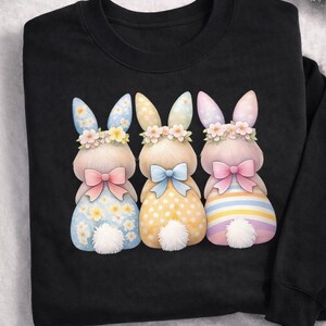 May include: Black sweatshirt featuring three watercolor bunny illustrations. Each bunny has a floral crown, bow, and patterned dress in blue, yellow, and pink. The bunnies have white fluffy tails.