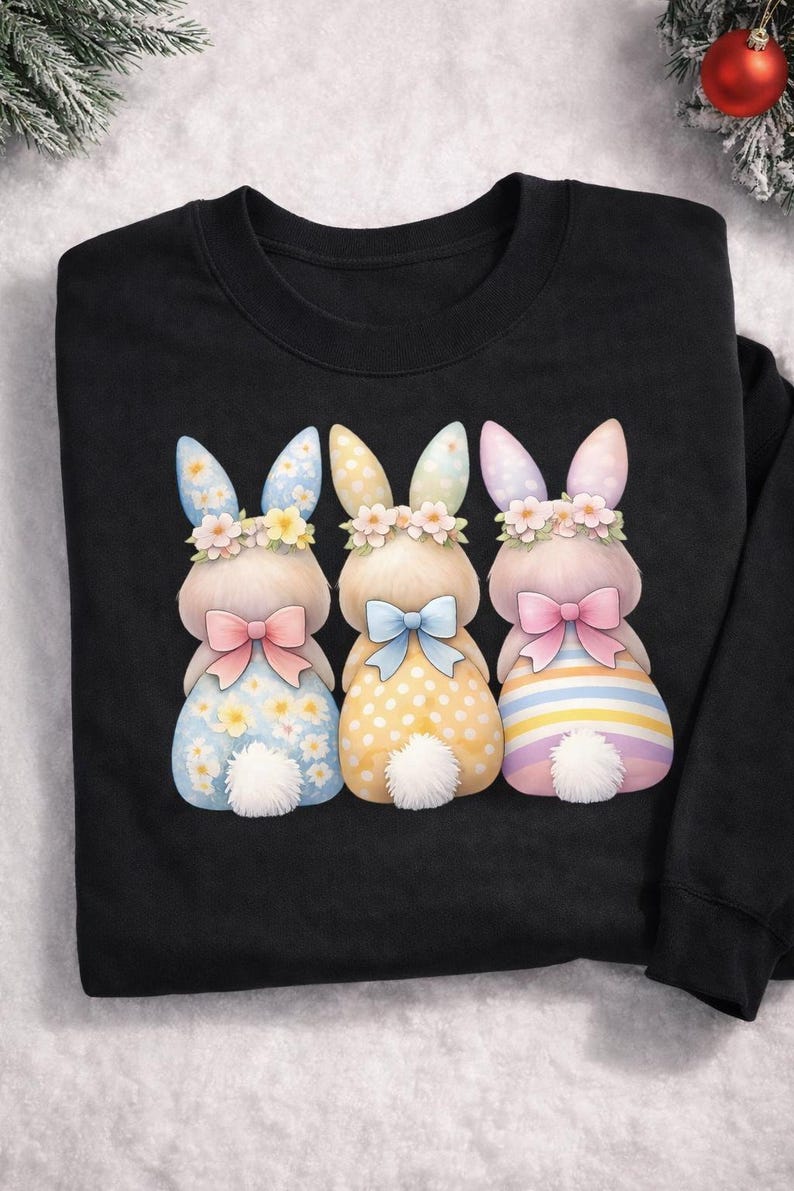 May include: Black sweatshirt featuring three watercolor bunny illustrations. Each bunny has a floral crown, bow, and patterned dress in blue, yellow, and pink. The bunnies have white fluffy tails.