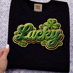 Może przedstawiać: Czarny t-shirt z napisem "Lucky" w zielonej, teksturowanej czcionce ze złotym obramowaniem. Zielone koniczyny są wkomponowane w projekt. Koszulka ma metkę z napisem "Comfort Colors".