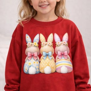 May include: A red sweatshirt featuring three cartoon bunnies with floral crowns and bows. The bunnies have patterned dresses in blue, yellow, and pink. The sweatshirt is a solid red color.