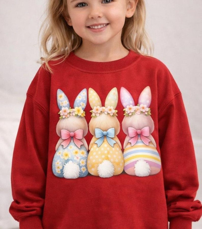 May include: A red sweatshirt featuring three cartoon bunnies with floral crowns and bows. The bunnies have patterned dresses in blue, yellow, and pink. The sweatshirt is a solid red color.