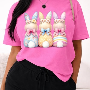 May include: A bright pink t-shirt featuring three cartoon bunny characters with floral crowns and bow ties. The bunnies have patterned bodies in blue, yellow, and striped designs. The shirt is paired with black shorts.