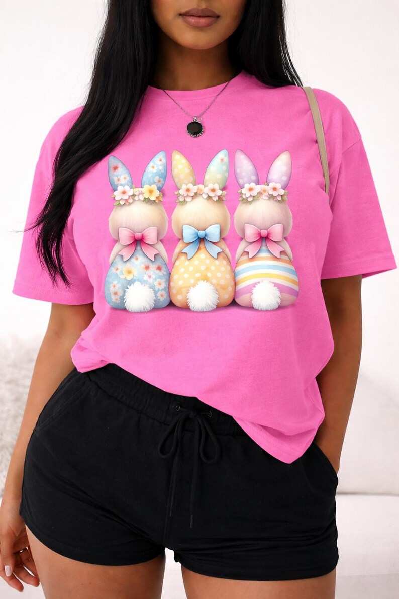 May include: A bright pink t-shirt featuring three cartoon bunny characters with floral crowns and bow ties. The bunnies have patterned bodies in blue, yellow, and striped designs. The shirt is paired with black shorts.