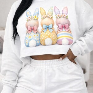 May include: White cropped sweatshirt and shorts set. The sweatshirt features a graphic of three Easter bunnies with floral crowns, bows, and patterned bodies in blue, yellow, and pink. The shorts are solid white.