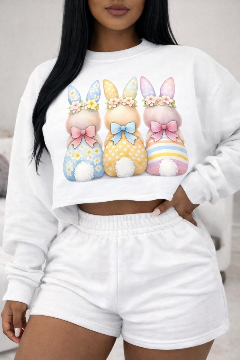 May include: White cropped sweatshirt and shorts set. The sweatshirt features a graphic of three Easter bunnies with floral crowns, bows, and patterned bodies in blue, yellow, and pink. The shorts are solid white.