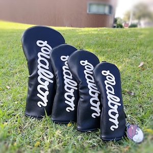 Puede incluir: Un juego de cuatro fundas negras para palos de golf con texto blanco en cursiva "Malbon". Las fundas están dispuestas sobre hierba. Es visible una pequeña etiqueta con un personaje de dibujos animados.