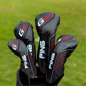Puede incluir: Un juego de fundas negras para palos de golf con el texto "G410" en rojo y blanco. Las fundas tienen un diseño texturizado con costuras rojas y la marca "PING" en blanco. El fondo es verde borroso.