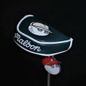 Puede incluir: Funda para la cabeza del palo de golf verde oscuro con ribete blanco y la palabra "Malbon" en escritura blanca. La funda presenta una bola de golf de dibujos animados con un sombrero. Una bola de golf más pequeña con un sombrero rojo cuelga debajo.