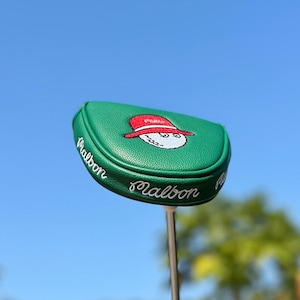 May include: A green golf club head cover with the word "Malbon" in white script. The cover features an embroidered design of a golf ball wearing a red hat. The background is a clear blue sky.