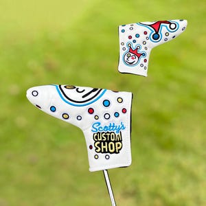 May include: Two white golf club head covers with colourful designs. One cover features a clown face and a crown, while the other displays the text "Scotty's Custom Shop" in blue and yellow. Both covers have a dotted pattern.